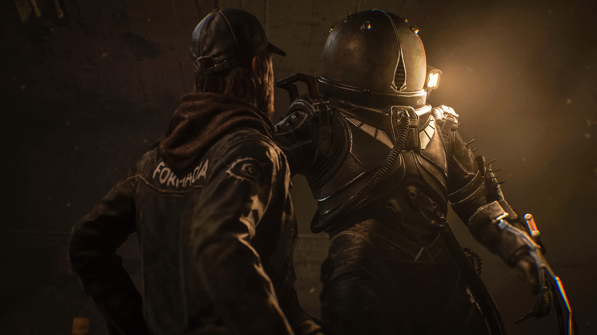 Two figures stand inside a dim room, one in a hood and one in a helmet.