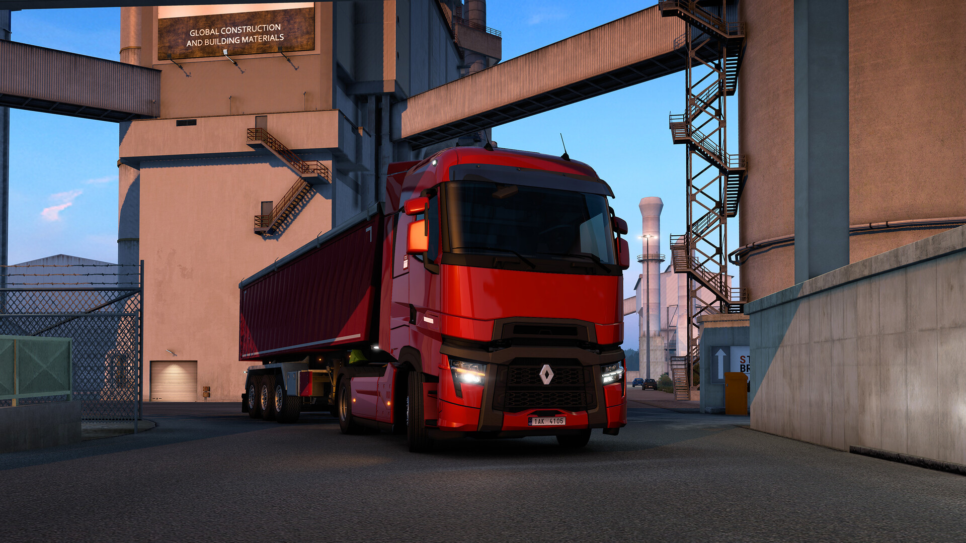 A red lorry carries a container beside an industrial loading site.