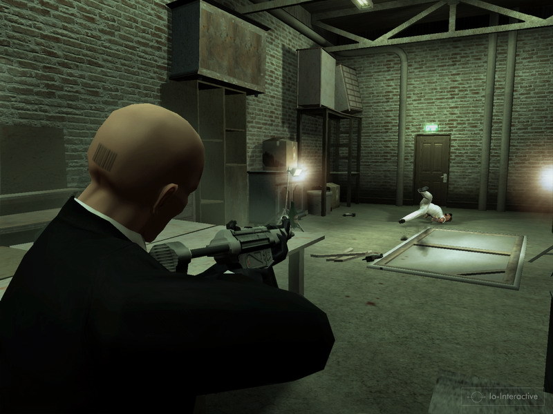 Agent 47 aims a weapon into a dim industrial room.
