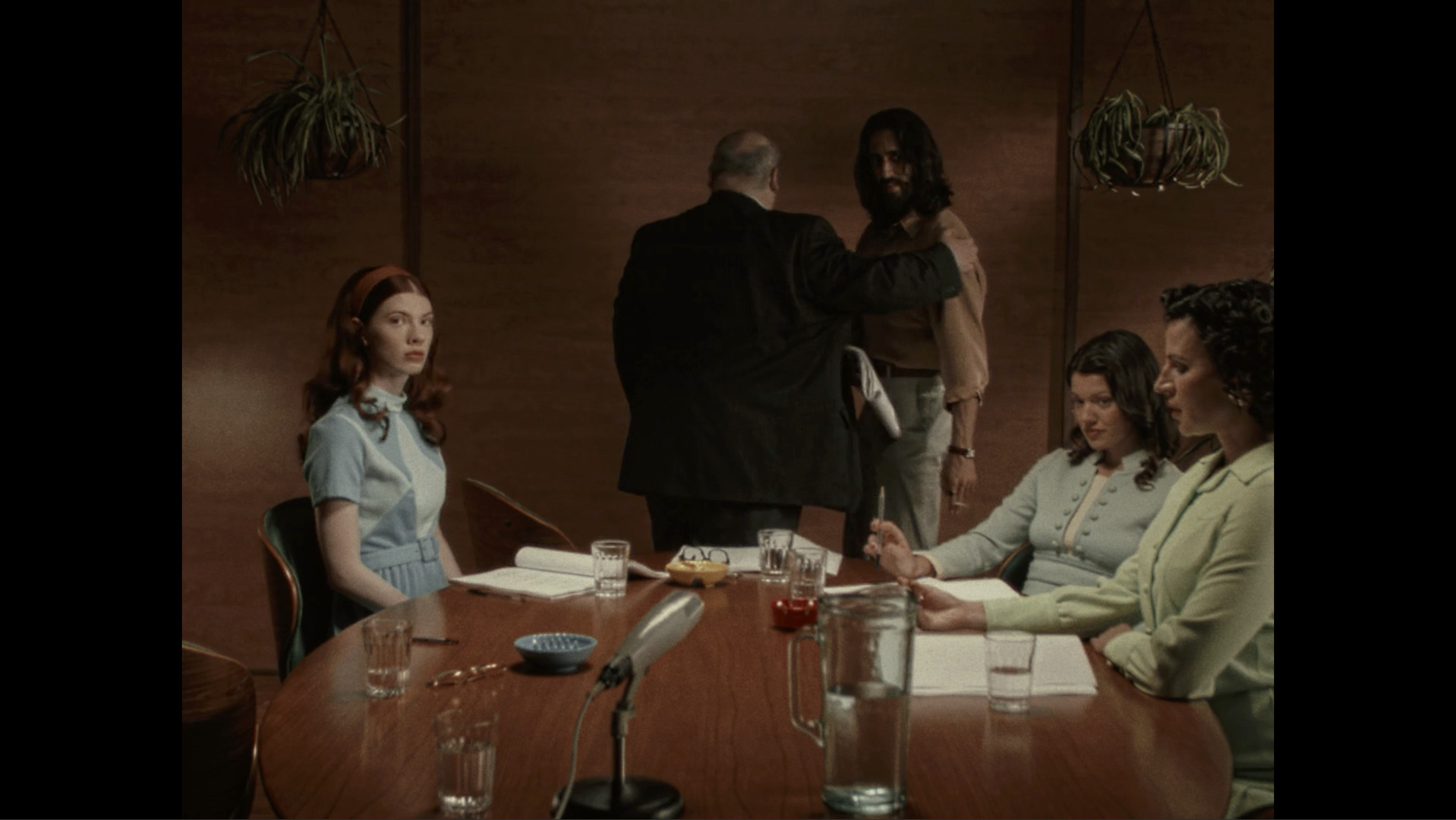 Actors sit around a table in a filmed scene.