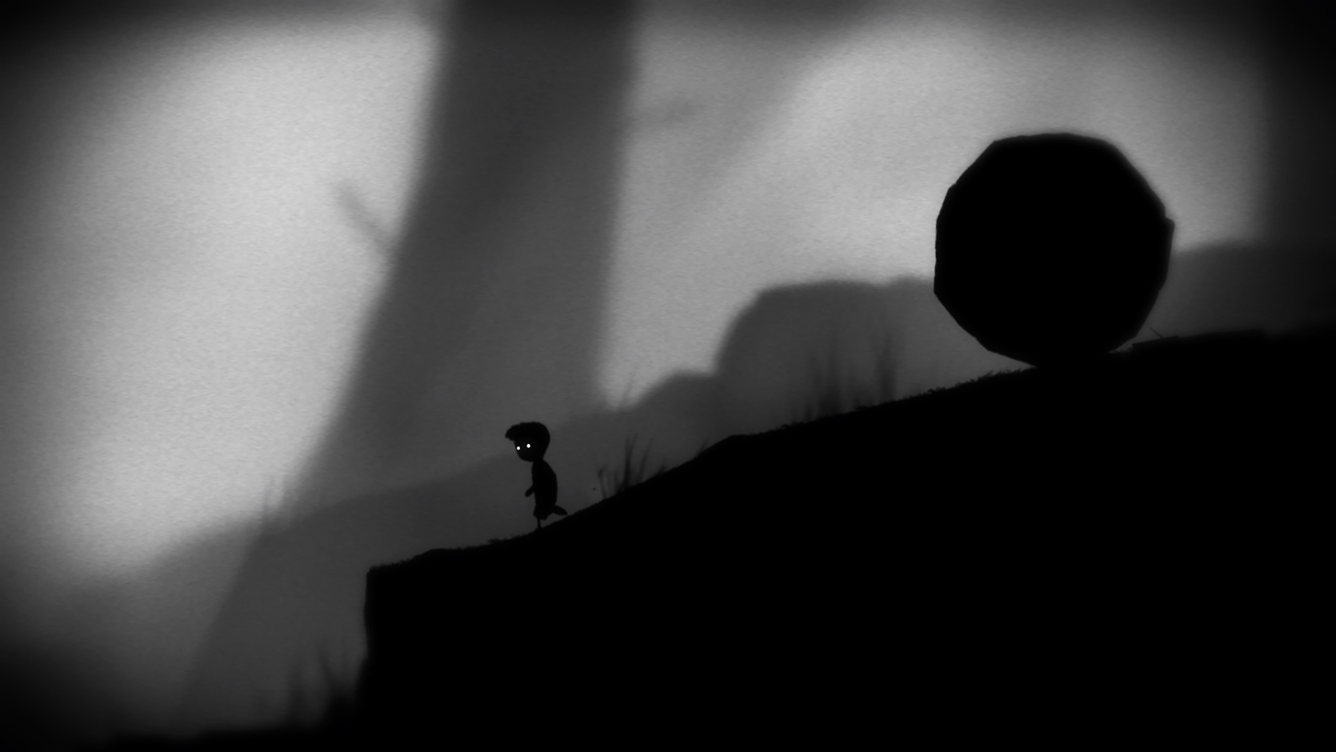 The boy stands beneath a large rolling boulder on a slope.